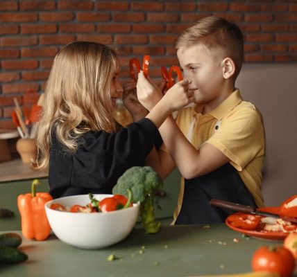 Playful boy and girl wearing apron preparing healthy vegetarian salad together at table with ingredients and bowls while looking at each other through slices of pepper