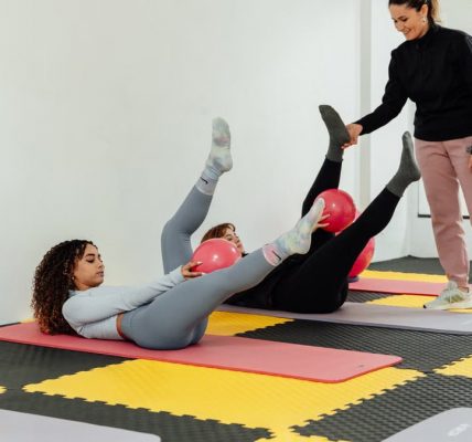 Women engaged in pilates class using pink balls and yoga mats indoors at a gym.